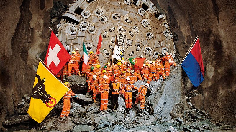 Das Baustellenpersonal steht fahnenschwenkend vor der Tunnelbohrmaschine nach dem finalen Durchbruch am Gotthard-Basistunnel. 