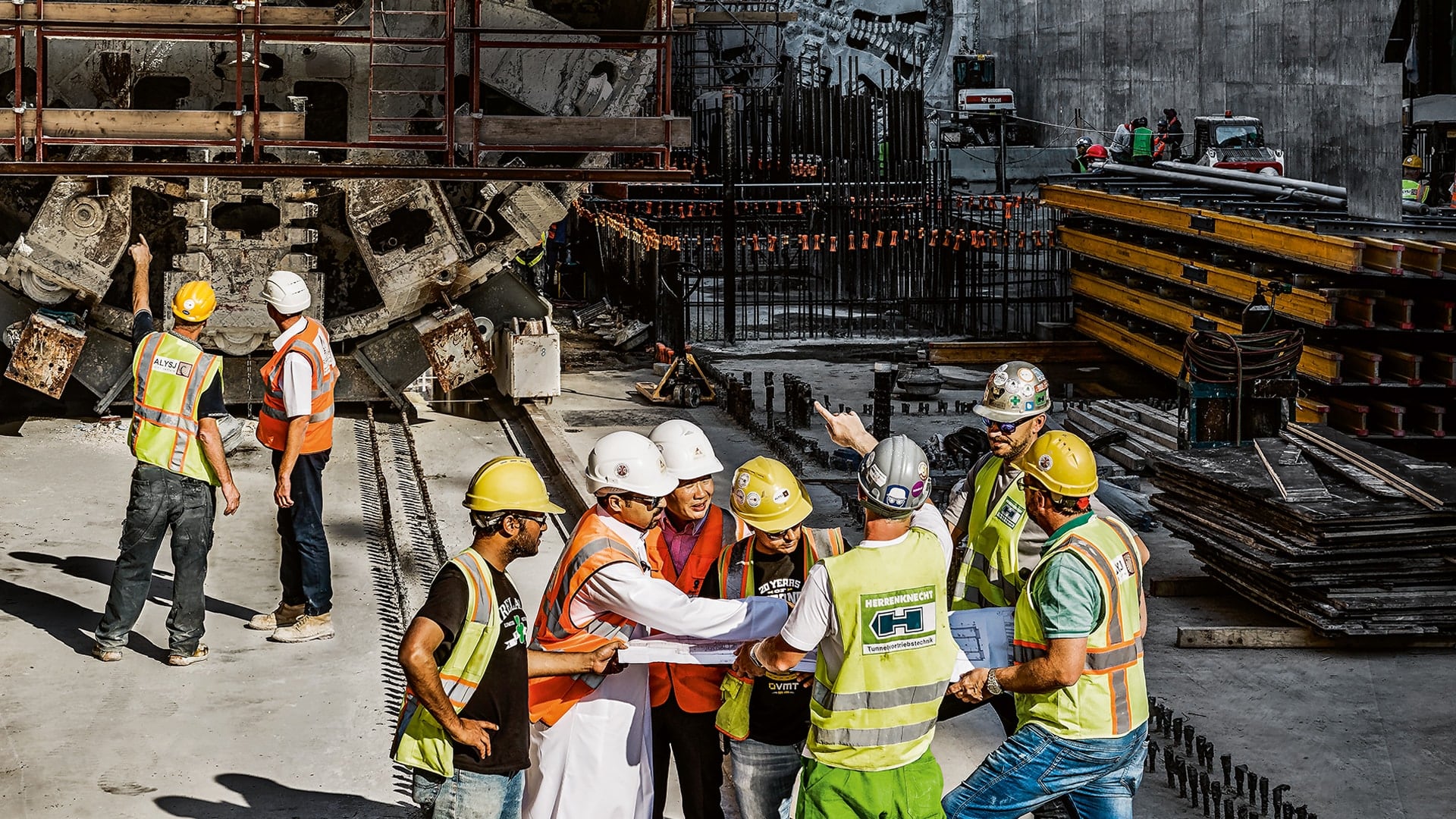 In the foreground, the construction site personnel are discussing a plan in their hands and in the background you can see other employees with two tunnel boring machines