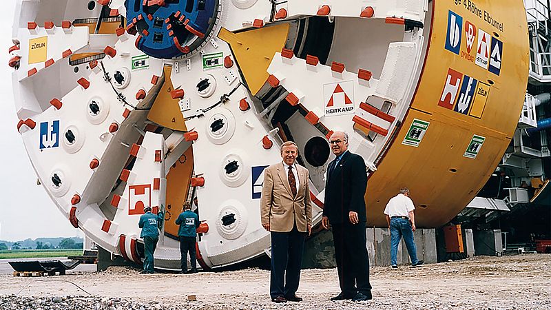 Two men stand in front of a yellow and white tunnel boring machine for the construction of the Elbe Tunnel.
