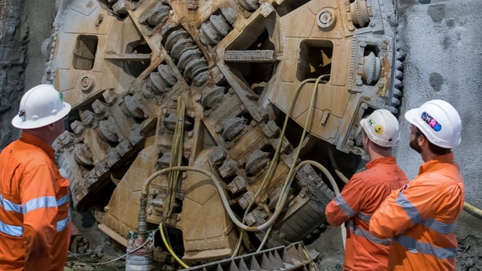 Drei Baustellenmitarbeiter betrachten eine Tunnelbohrmaschine einer Metro-Baustelle